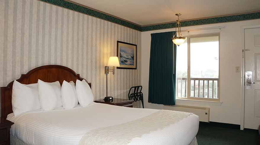 Photo showing queen bed, night stand with lamp and alarm clock. Large viewing window and luggage rack.