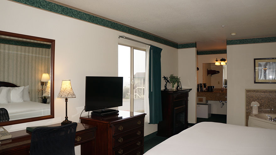 European King Size Bed guest room showing photo of partial bed, jacuzzi, chest of drawers with flat screen tv, writing desk, lamp, bathroom.